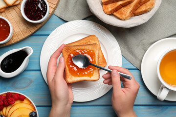 Woman spreading apricot jam onto tasty toast at light blue wooden table, top view