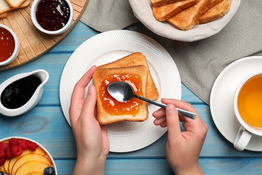 Woman spreading apricot jam onto tasty toast at light blue wooden table, top view