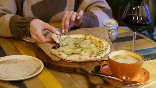 A woman's hand takes a piece of hot pizza with pear and cheese, sitting in a cafe in the evening.