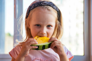 Funny little toddler girl eating yellow watermelon close-up at window background, looking at camera. Portrait blonde child with watermelon at home. Concept of healthy food, summertime. Copy text space