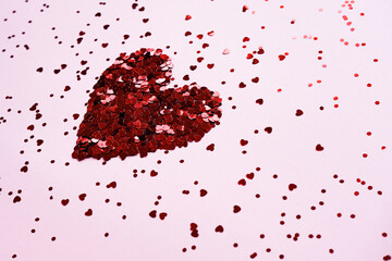 Heart made of shiny red small decorative hearts on a pink background strewn with sparkles. Side view, space for text. Valentine's Day card.