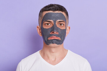 Portrait of handsome brunet man with sad expression, wears nourishing homemade mud mask for reducing dark dotes, wears casual white t shirt, isolated over purple background.