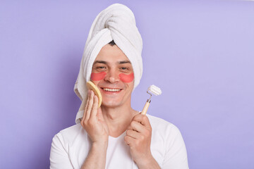 Indoor shot of smiling delighted man wearing white T-shirt and being wrapped in towel standing isolated over purple background, massaging his face with roller.