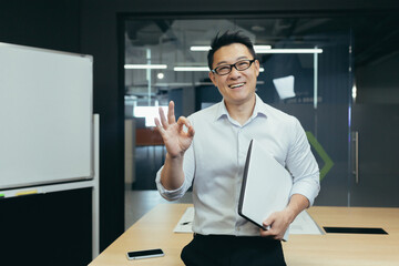 Portrait of a young handsome Asian student, intern, assistant. He stands with a folder in the office, wears glasses, smiles, points with his fingers
