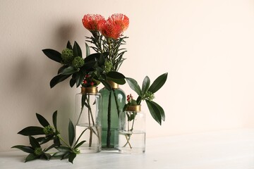 Vases with beautiful flowers and branches on white wooden table near beige wall. Space for text