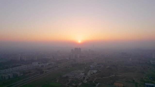 aerial drone shot of flying towards colorful sunset with skyscrapers in foreground and busy street and barren feilds below in gurgaon delhi jaipur showing development and growth