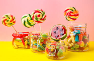 Tasty colorful candies in glass jars on yellow table against pink background