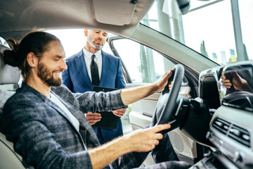 Satisfied male customer sitting at the wheel of new car serious dealer explaining automobile characteristics buying cars.