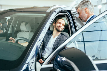 Portrait of handsome young man taking luxury car for test drive, sitting inside with mature bearded professional salesman standing near car in showroom.