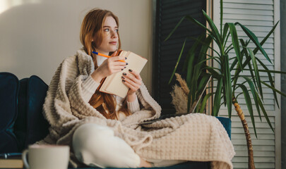 Thoughtful and cozy red-haired woman sitting with her legs crossed and taking notes on notebook....
