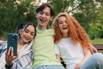 Cheerful smiling group of friends sitting on a bench in the city park and having fun while posing for selfie together. Holiday life moment with technology