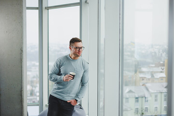 Fototapeta premium Smiling man in casual clothes standing near window with coffee in modern work area with big window during daytime.