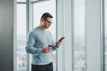 Fototapeta na wymiar Smiling man in casual clothes standing near window with coffee and looking at smartphone in modern workspace with big window during daytime