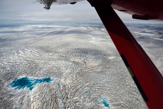 A large pool of water remains on the surface of the ice cap as the Twin Otter aeroplane flies over the ice. Surface structure of the glacier is apparent. Photograph taken through the window of the Twi