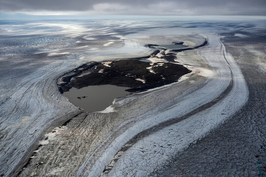 A giant glacier flows down from off the ice cap towards the coast exposing a circular area of rock appearing in the middle. Photograph taken through the window of the Twin Otter aeroplane enroute to C