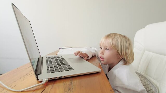 Caucasian 5 years old boy in his fathers's shirt and tie is watching something on the laptop screen. A boy is using a laptop at the desk to atch videos