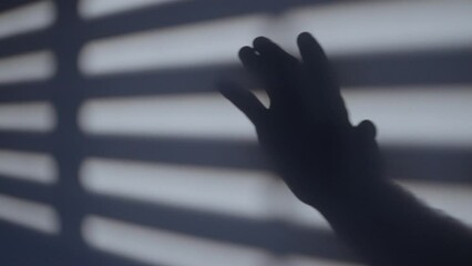 Hard window blind shadows cast on wall with hand shadow waving in dramatic scene. Freedom and jail context. Blue colored scene. Stuck in small room or cell. Waving goodbye or hello