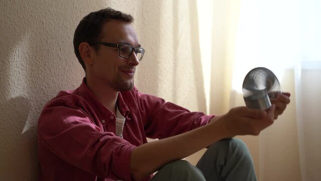 Young relaxed man sitting on floor playing with anti-stress metal spring toy for calmness and smiling, distracting from negative thoughts. Fun home activities, stress relief concept