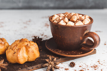 Hot chocolate drink with marshmallows in a cup and sweet bakery eclair dessert sprinkled with cocoa powder, cinnamon stick and star anise on white table