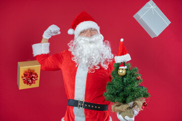 Portrait von einem verkleideten Weihnachtsmann mit Geschenk und Weihnachtsbaum, isoliert auf roten Hintergrund im Studio 
