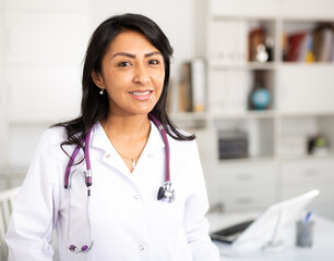 Portrait of confident smiling female doctor in white lab coat standing in modern medical office