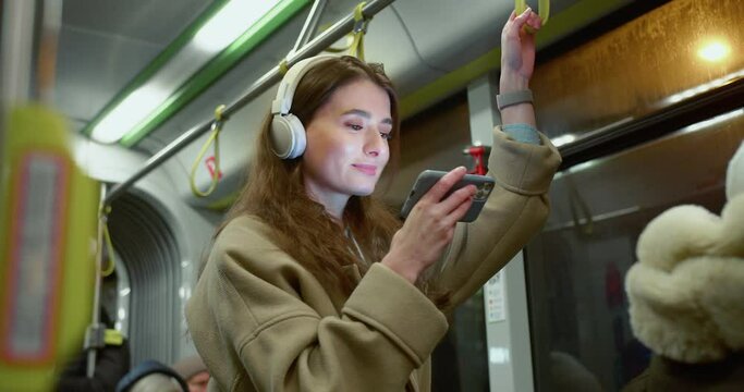 Side-view portrait of attractive happy caucasian female using public transport standing holding handrail watching videos on smartphone and wireless headphones. Charming young woman travelling by bus.