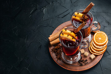 Two glasses of hot mulled wine stand on a dark background. A tray with ingredients for mulled wine - cinnamon, star anise, citrus fruits. Place for text.