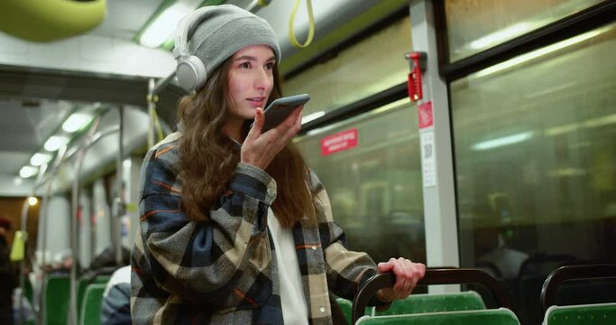 Portrait young beautiful woman hipster recording and listening to voice messages using smartphone online. Cute happy female riding bus smiling wearing shirt, beanie and headphones. Public transport.