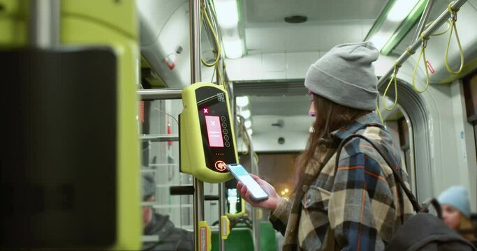 Back-view of young woman in shirt and beanie paying online with smartphone in public transport. Attractive modern caucasian female riding a bus buying tickets by using NFC technologies.