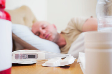 Pills and electric thermometer in focus. Bald sick man out of focus in the background. Flu and winter season infection concept.