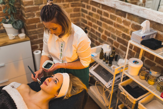 Cosmetologist applying black mask on the face of a beautiful blond woman at the spa, skincare concept. High quality photo