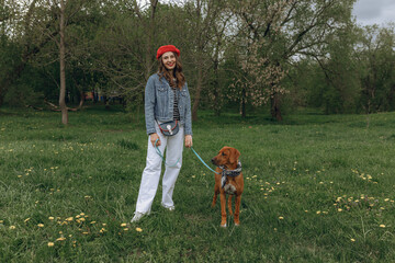 Full body of content woman standing with Rhodesian Ridgeback puppy on leash on lawn in park and looking at camera 