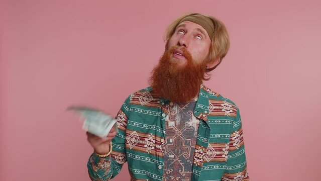Rich pleased boss bearded hipster man waving money dollar cash banknotes bills like a fan, success business career, lottery winner, big income, wealth. Young hippie guy boy on pink studio background