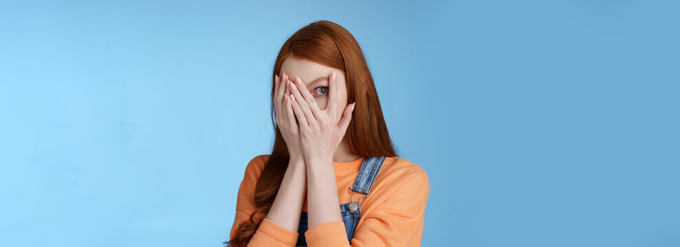 Not Peeking Promise. Charming Intrigued Cute Redhead Teenage Girl Hiding Face Cover Eyes Palms Look Through Fingers Check Out Gift Anticipating Something Interesting Standing Curious Blue Background