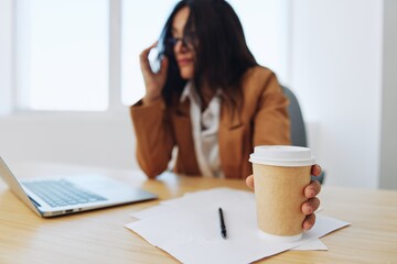 Woman business works in the office at the desk in the office in a beige suit, glasses with a cup of coffee, autumn mood, tiredness coffee break