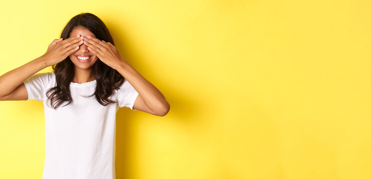 Image Of Excited African-american Girl Waiting For Surprise, Smiling And Covering Eyes With Hands, Standing Over Yellow Background