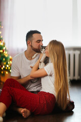 Cozy Christmas lovers. Abandoned kitten from shelter mongrel kitten at home. Cute traditional couple in pajamas with kitten on floor in living room by Christmas tree.