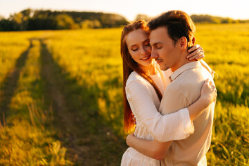 Fototapeta premium Medium shot portrait of young couple in love with closed eyes standing embracing together on green meadow in summer evening, during sunset with soft sunlight. Concept of romantic date love outdoors.