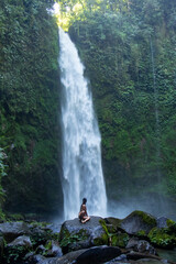 Young Asian girl on large rock looking up at massive waterfall in rainforest - Bali, Indonesia