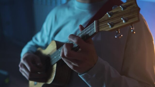 An adult smiling man plays the ukulele in the light neon dark room. A strong and blond guy sits and plays a small guitar in atmosphere place. A happy person plays a musical instrument.