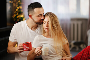 Young man tender kiss woman drink coffee from mugs hugging on living room floor. Cute European couple with cocoa in their pajamas in living room by Christmas tree. Cozy good Christmas morning.