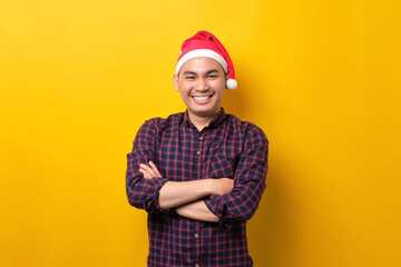 Smiling young Asian man in Santa hat standing with crossed arms, looking at camera over yellow studio background. Happy New Year 2023 celebration merry holiday concept