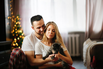 Cozy Christmas lovers. Cute traditional couple in pajamas with kitten on floor in living room by Christmas tree. Kitten of Maine Coon breed in arms of owners.