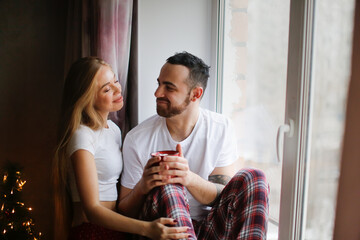 Portrait of young man and woman talking on window sill near window. Copy space. Cute couple with cocoa in pajamas in real room. Cozy good Christmas morning.