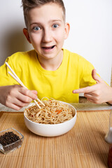 The boy points in surprise at the noodles in the white bowl, a dish of Asian cuisine. Close-up portrait.