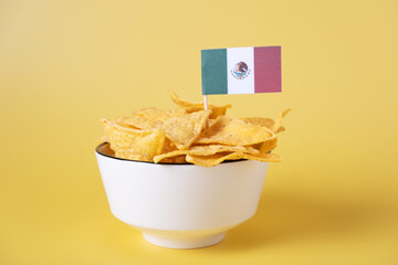 Corn nacho chips in a white bowl on a yellow background with the flag of Mexico.