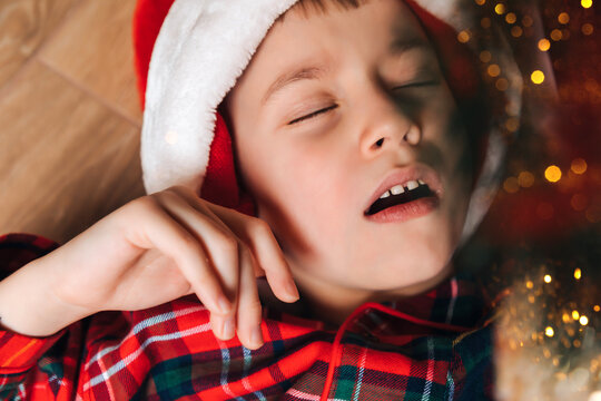 A sleeping boy in red-green pajamas and a Santa hat lies on the floor. Christmas at home. Top view