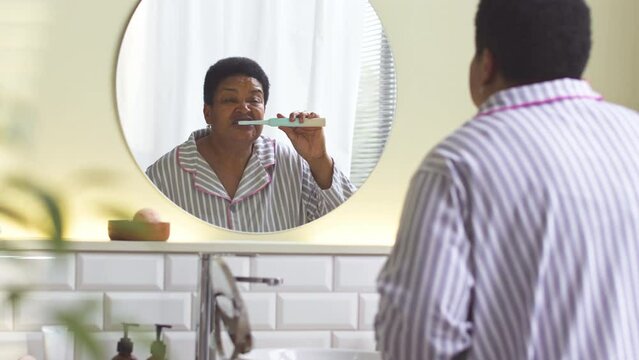 Middle aged Black woman in striped pajamas standing in front of round mirror in bathroom brushing her teeth in morning