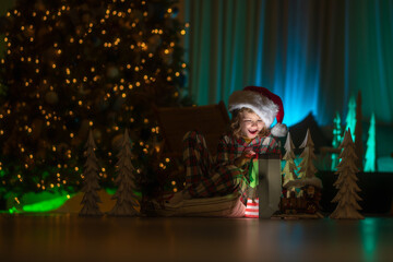Child opening presents on Xmas eve. Kid in Christmas pajama enjoying winter holiday evening at home near the night Christmas tree.