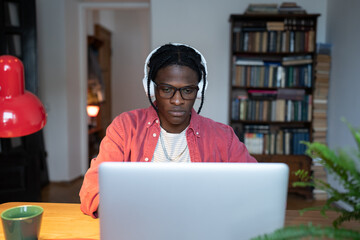 Focused concentrated young African man freelancer wearing headphones listening to music while working remotely from home, using laptop at remote workplace. Distance Learning, freelance concept